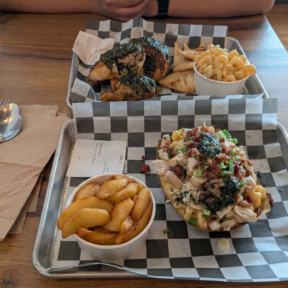 Two restaurant trays hold loaded macaroni and cheese, fries, chicken wings, pita, and dipping cups on a wooden table.