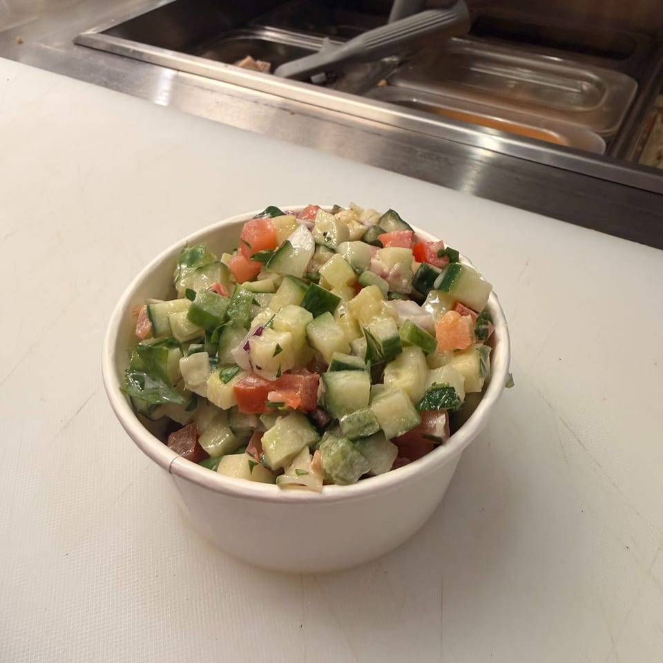 Bowl of chopped cucumber, tomato, onion, and herbs at a restaurant counter with stainless-steel food trays.