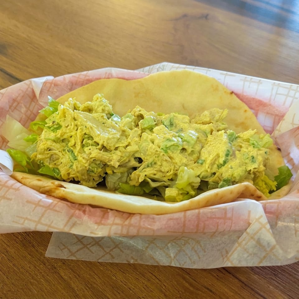 Chicken salad piled in warm flatbread with lettuce, served in patterned paper on a wooden restaurant table.