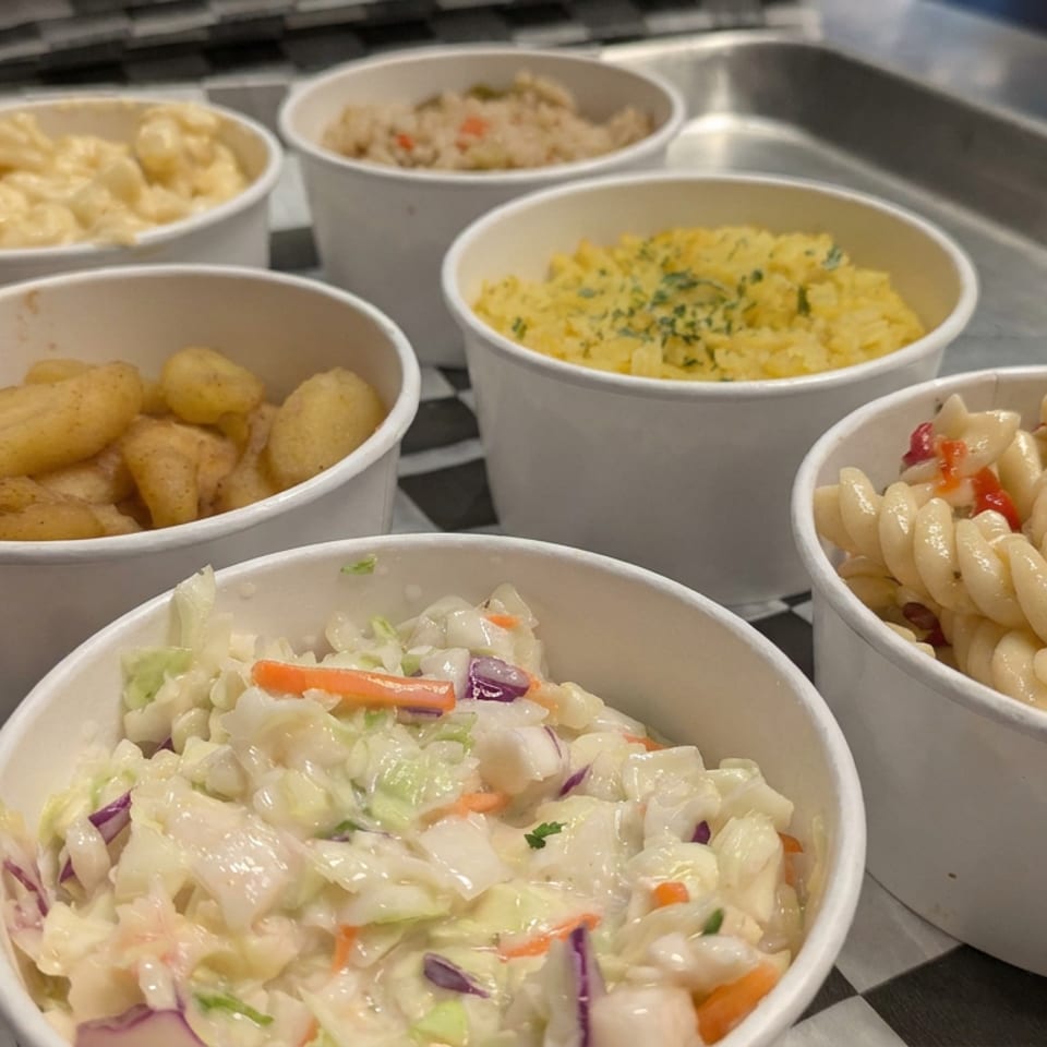 Six white bowls hold assorted side dishes, including macaroni salad, rotini pasta salad, potatoes, mashed potatoes, rice, and macaroni and cheese.