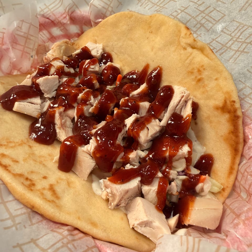 Flatbread topped with chopped chicken, vegetables, and glossy red sauce, served on patterned paper beside a food preparation counter.