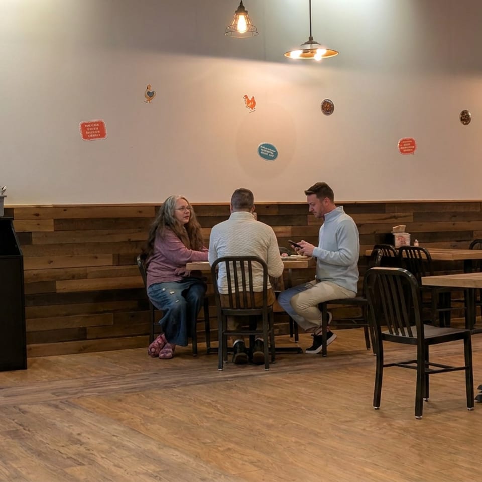 Warmly lit restaurant dining room with wooden walls, pendant lights, scattered tables, and three people seated together.