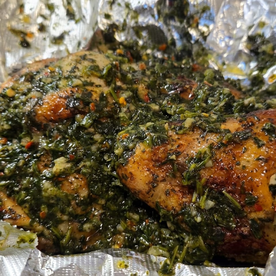 Grilled chicken pieces coated in a chunky green herb sauce, served wrapped in crinkled aluminum foil.